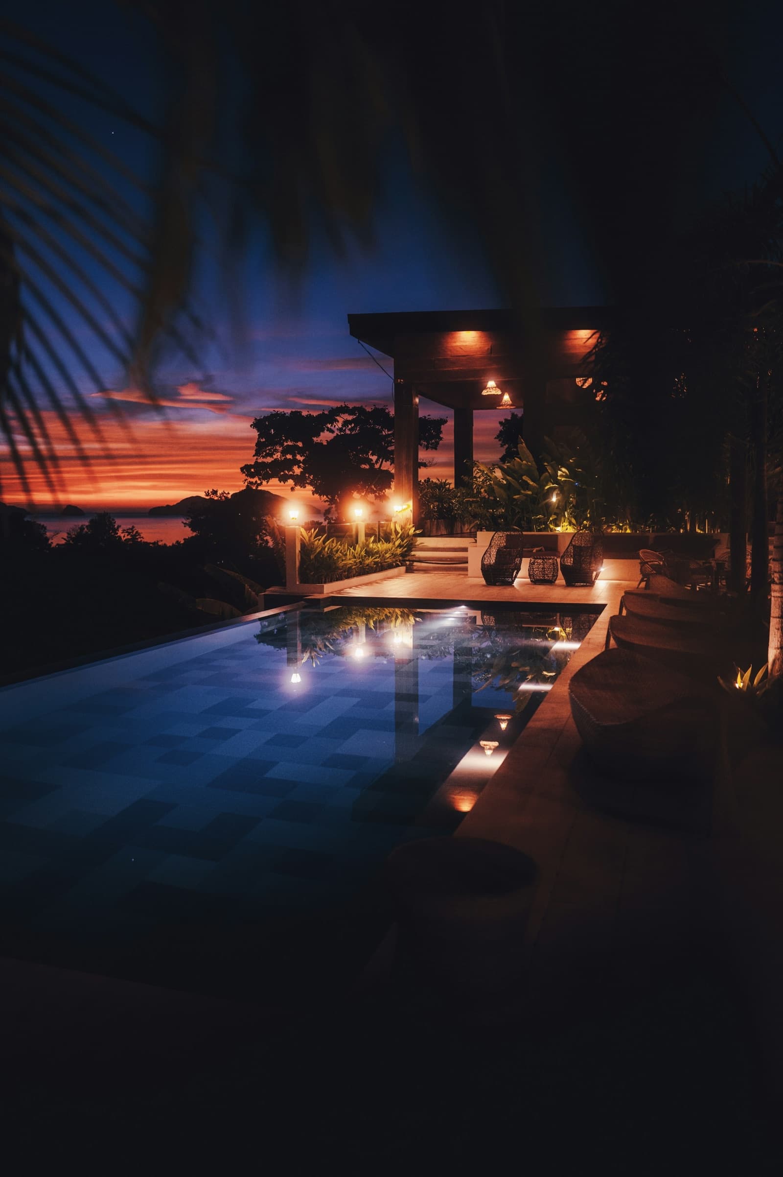 A private villa terrace with a lit pool at dusk, framed by palm leaves.
